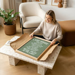 A woman is sitting on the floor in a living room, holding a framed sign with text on it.