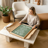 A woman is sitting on the floor in a living room, holding a framed sign with text on it.