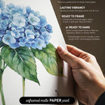 a hand holding a piece of paper with a close-up view of a watercolor painting of blue hydrangeas.