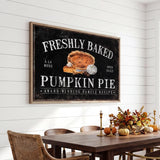 a framed sign that reads "FRESHLY BAKED PUMPKIN PIE" above a wooden dining table with chairs. The table is set with a vase of flowers and a candle, creating a cozy and inviting atmosphere.