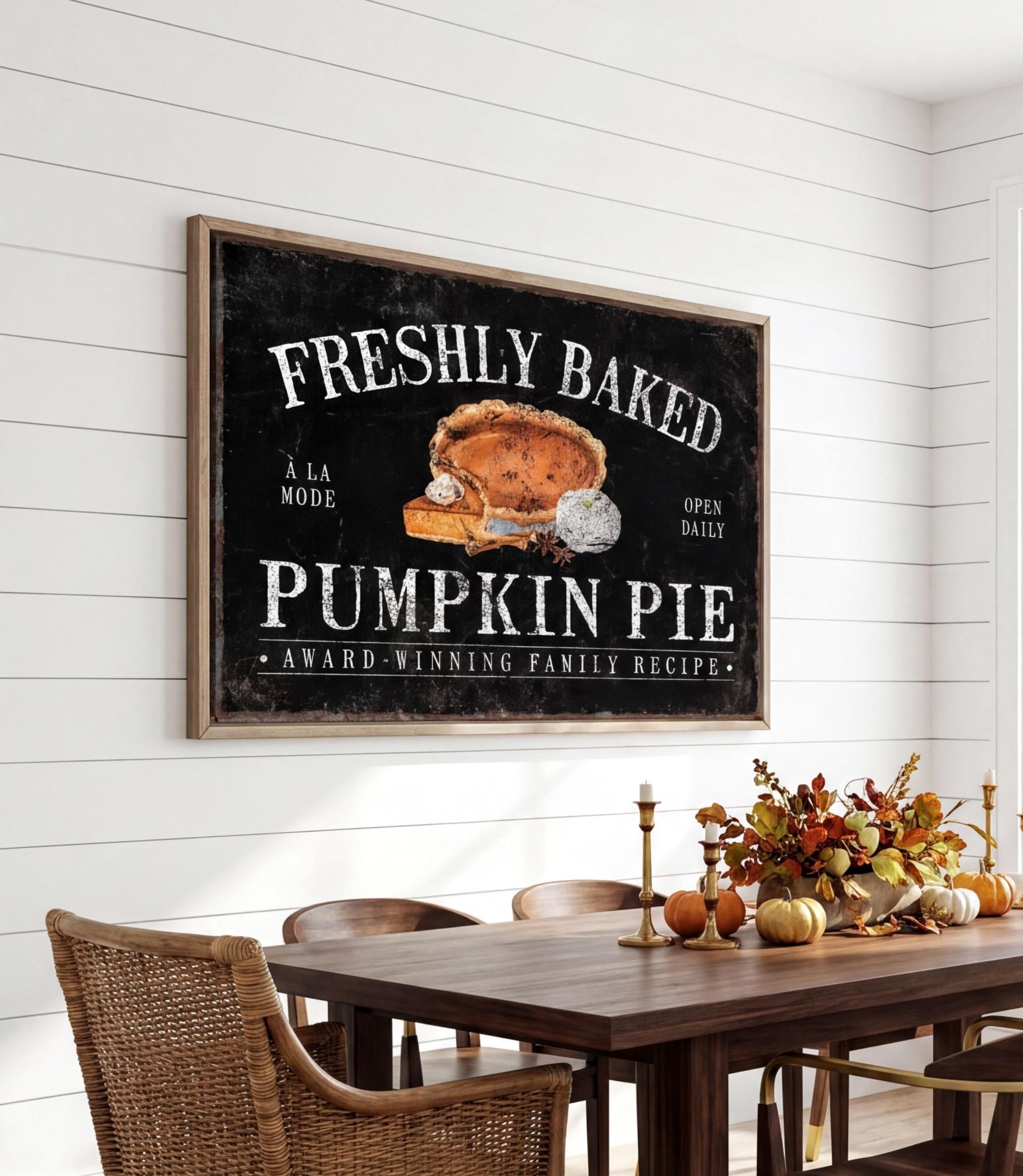 a framed sign that reads "FRESHLY BAKED PUMPKIN PIE" above a wooden dining table with chairs. The table is set with a vase of flowers and a candle, creating a cozy and inviting atmosphere.