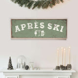 a white fireplace mantel decorated with various holiday items, including a green sign that says "APRES SKI", a glass dome, pine cones, and candles.
