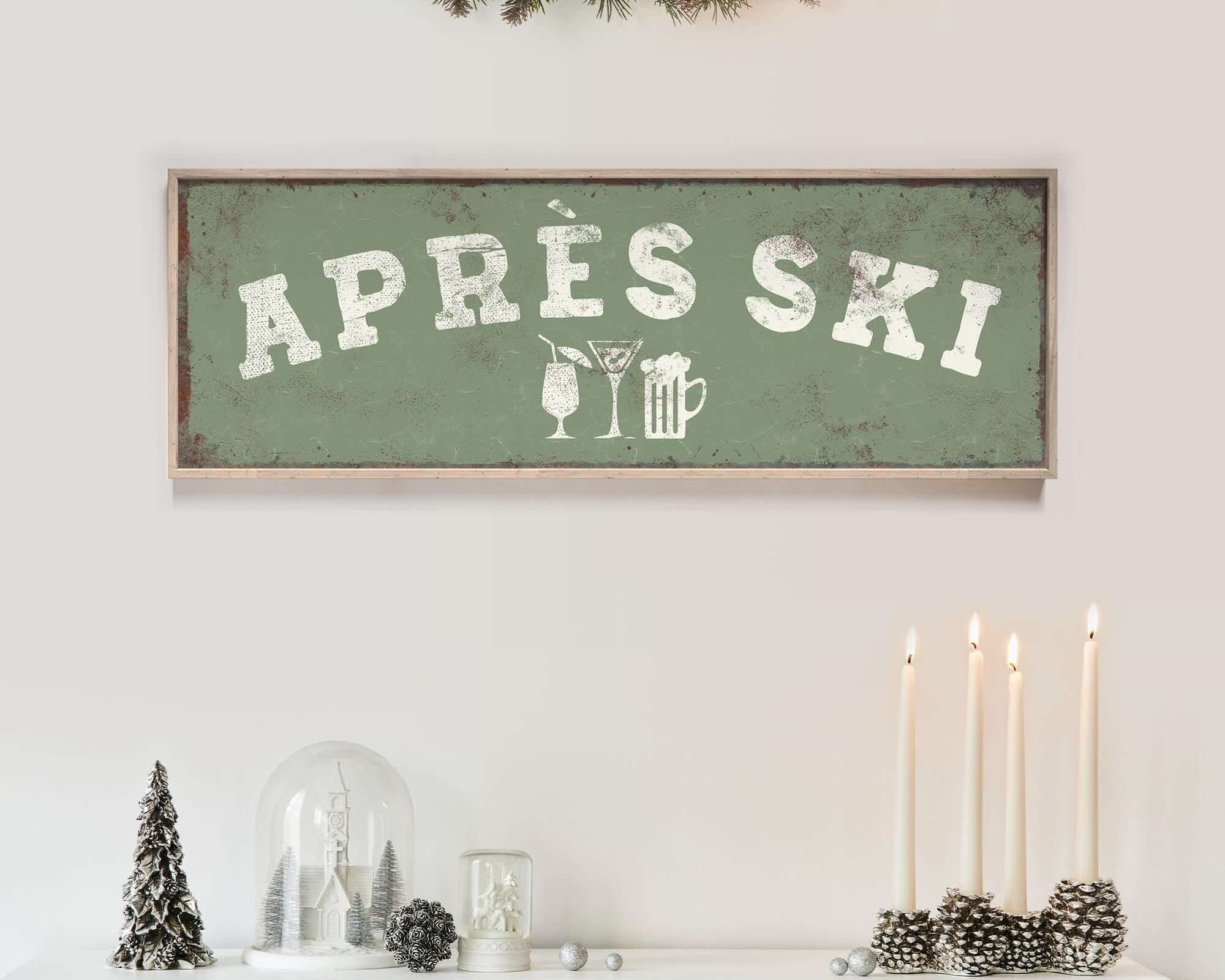 a white fireplace mantel decorated with various holiday items, including a green sign that says "APRES SKI", a glass dome, pine cones, and candles.