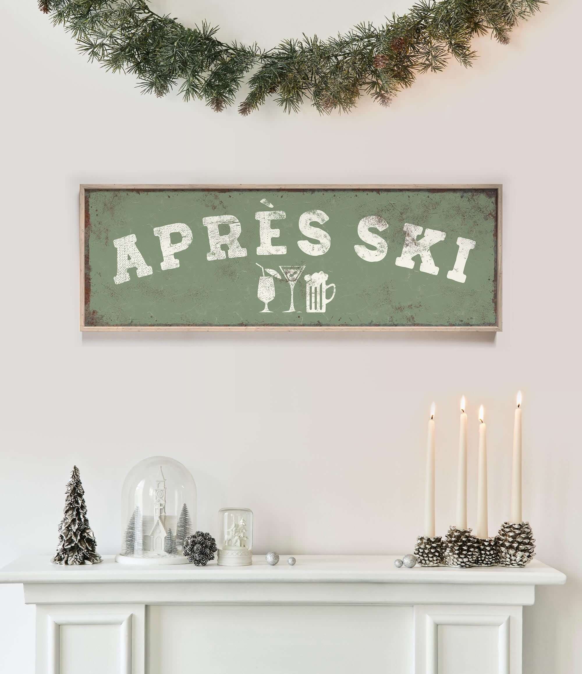 a white fireplace mantel decorated with various holiday items, including a green sign that says "APRES SKI", a glass dome, pine cones, and candles.