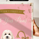 a hand holding a framed canvas print with a pink background and a white dog. The text on the canvas reads "Don't forget to wash your paws".