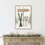 a framed picture of a ski sign with a winter scene below it, including a pair of skis and a small village. The picture is hung on a wall above a wooden console table with a wreath and candles on it.