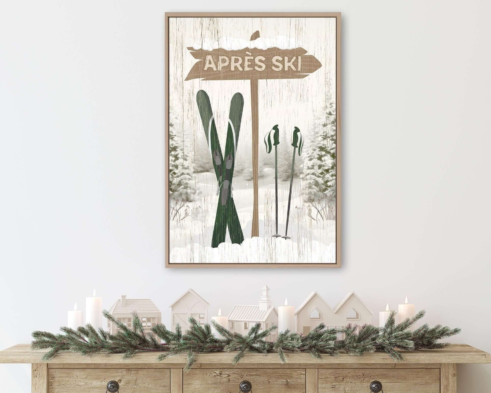 a framed picture of a ski sign with a winter scene below it, including a pair of skis and a small village. The picture is hung on a wall above a wooden console table with a wreath and candles on it.