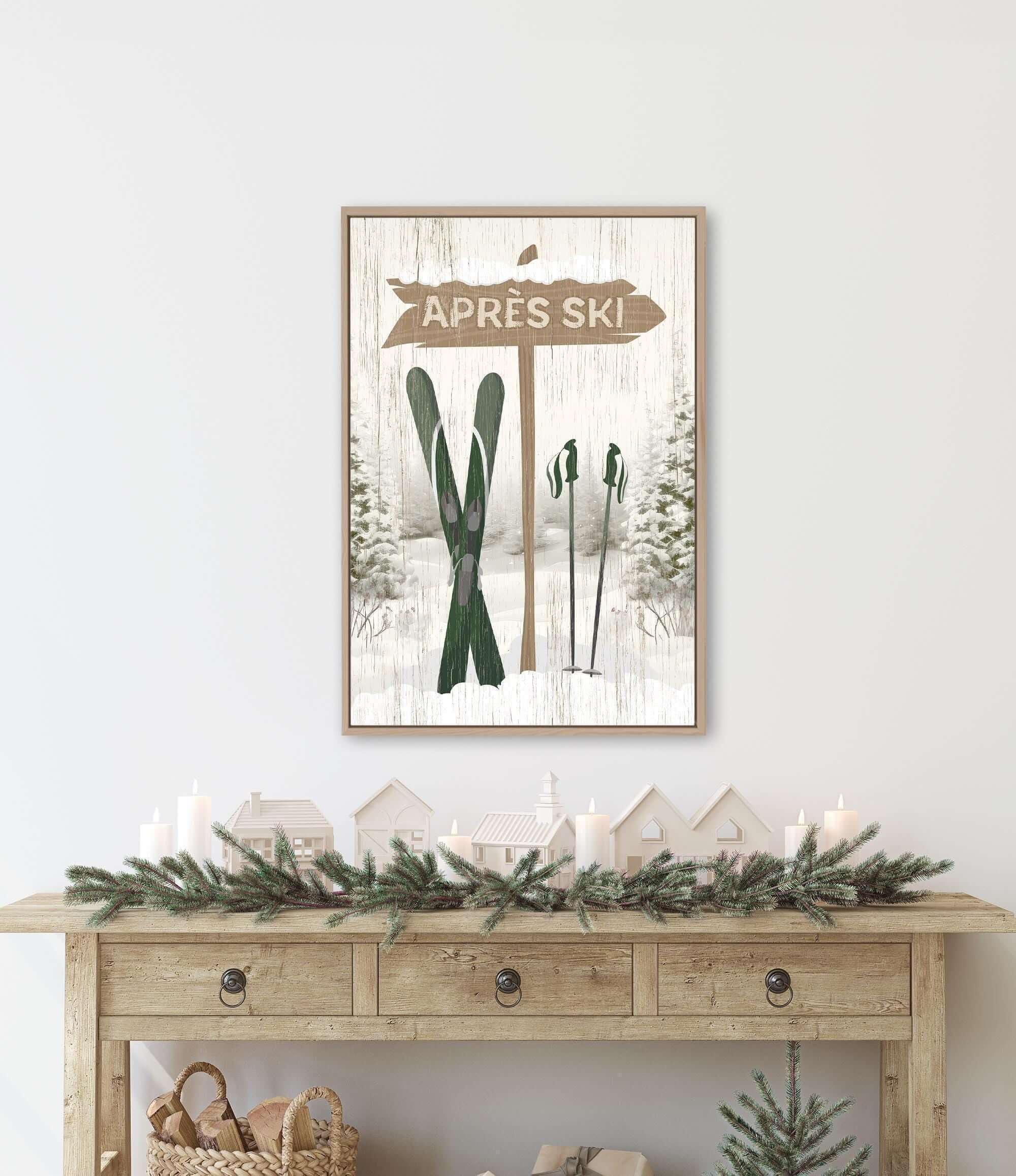 a framed picture of a ski sign with a winter scene below it, including a pair of skis and a small village. The picture is hung on a wall above a wooden console table with a wreath and candles on it.