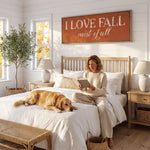 A woman sits on a bed in a cozy bedroom, reading a book, with a golden retriever dog resting on the bed beside her. The room has a rustic, country-style decor, with a large orange sign that reads "I LOVE FALL mist of fall" above the bed.