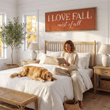 A woman sits on a bed in a cozy bedroom, reading a book, with a golden retriever dog resting on the bed beside her. The room has a rustic, country-style decor, with a large orange sign that reads "I LOVE FALL mist of fall" above the bed.