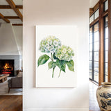 a large white canvas hanging on a wall, depicting a bouquet of white hydrangea flowers with green leaves. The canvas is positioned in the center of the wall, surrounded by a white frame. In the background, there is a fireplace and a large vase, creating a cozy and inviting atmosphere in the room.