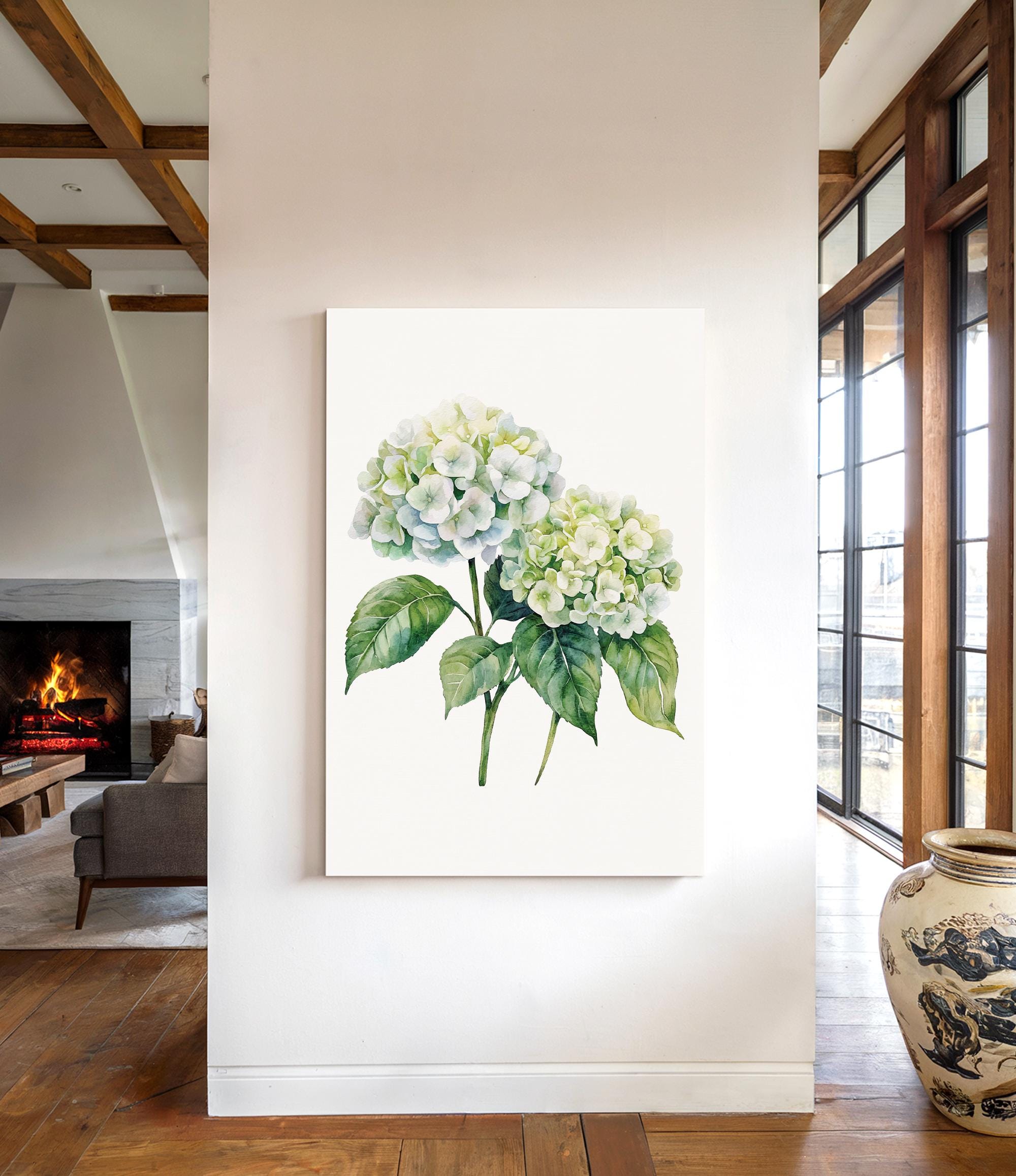 a large white canvas hanging on a wall, depicting a bouquet of white hydrangea flowers with green leaves. The canvas is positioned in the center of the wall, surrounded by a white frame. In the background, there is a fireplace and a large vase, creating a cozy and inviting atmosphere in the room.