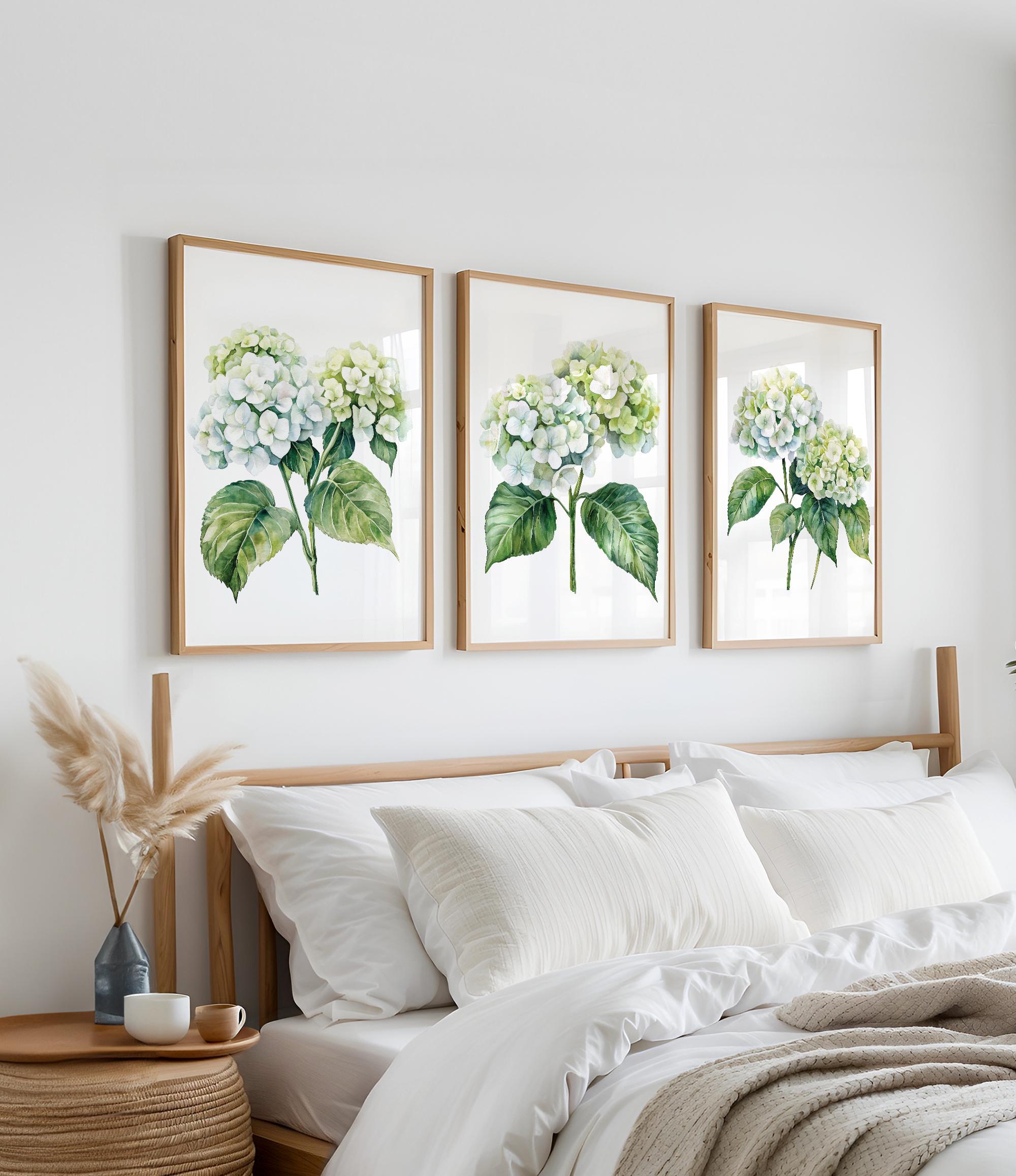 a bedroom with a bed, white bedding, and three framed watercolor paintings of white hydrangea flowers hanging on the wall above the bed.