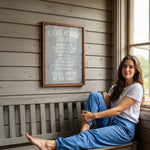 A woman sits on a bench in front of a sign that reads "LAKESHOUSE RULES" in a wooden cabin setting.