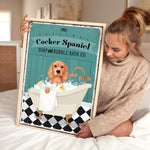 A woman is sitting on a bed holding a framed poster of a Cocker Spaniel in a bathtub, with a towel and bubbles around it.