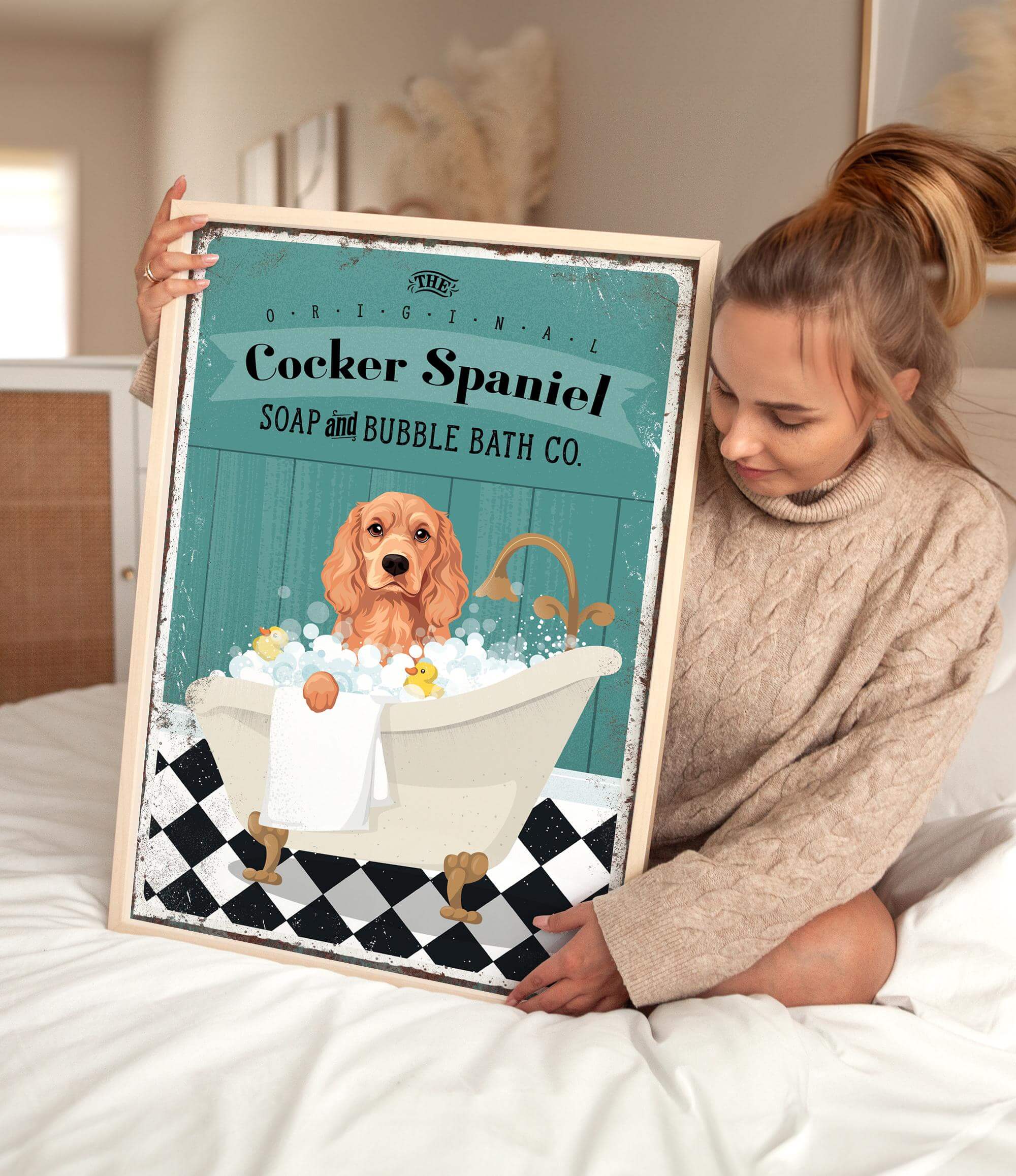 A woman is sitting on a bed holding a framed poster of a Cocker Spaniel in a bathtub, with a towel and bubbles around it.