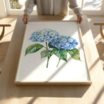 A person is holding up a framed painting of blue hydrangea flowers on a wooden table.