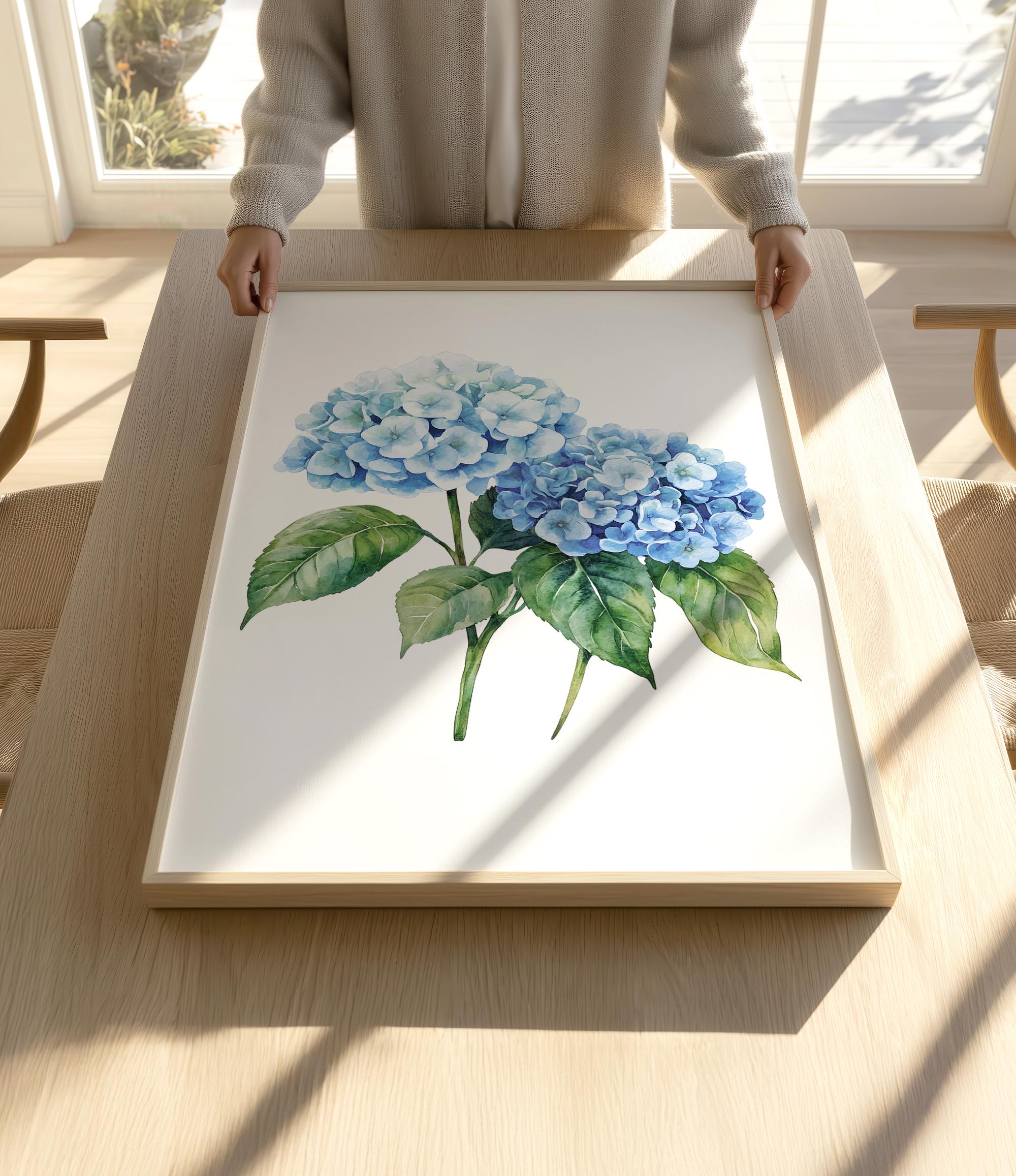 A person is holding up a framed painting of blue hydrangea flowers on a wooden table.