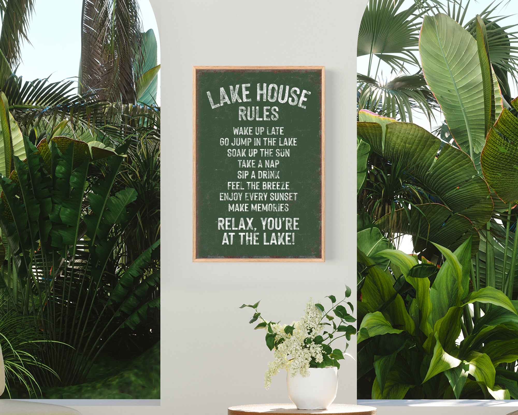 a sign with rules for a lake house, surrounded by lush tropical plants and a wooden table with a vase of white flowers.
