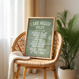 A framed sign on a wicker chair, displaying the rules of a lake house, with a potted plant in the background.