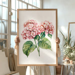 A person is holding up a framed painting of pink hydrangea flowers in a room with various potted plants and art supplies.