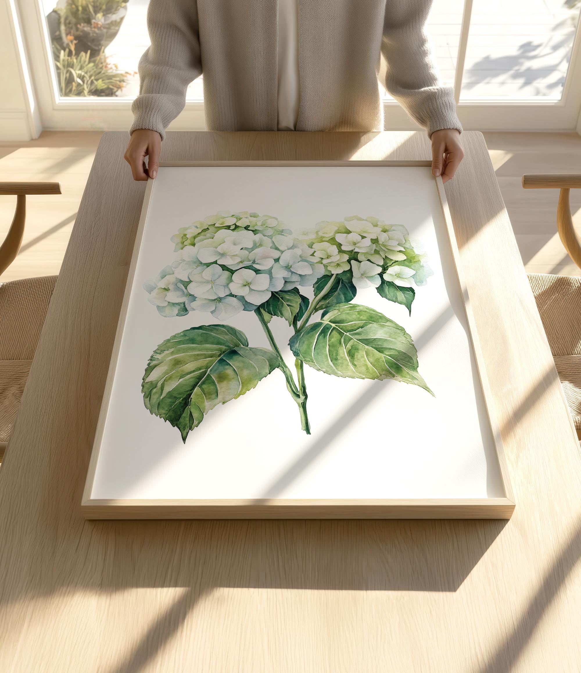 A person is holding a framed print of a watercolor painting of white hydrangea flowers on a wooden table.