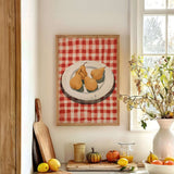 a kitchen counter with a framed painting of pears on a red and white checkered tablecloth, a wooden cutting board, a vase of flowers, and various fruits and vegetables on the counter.