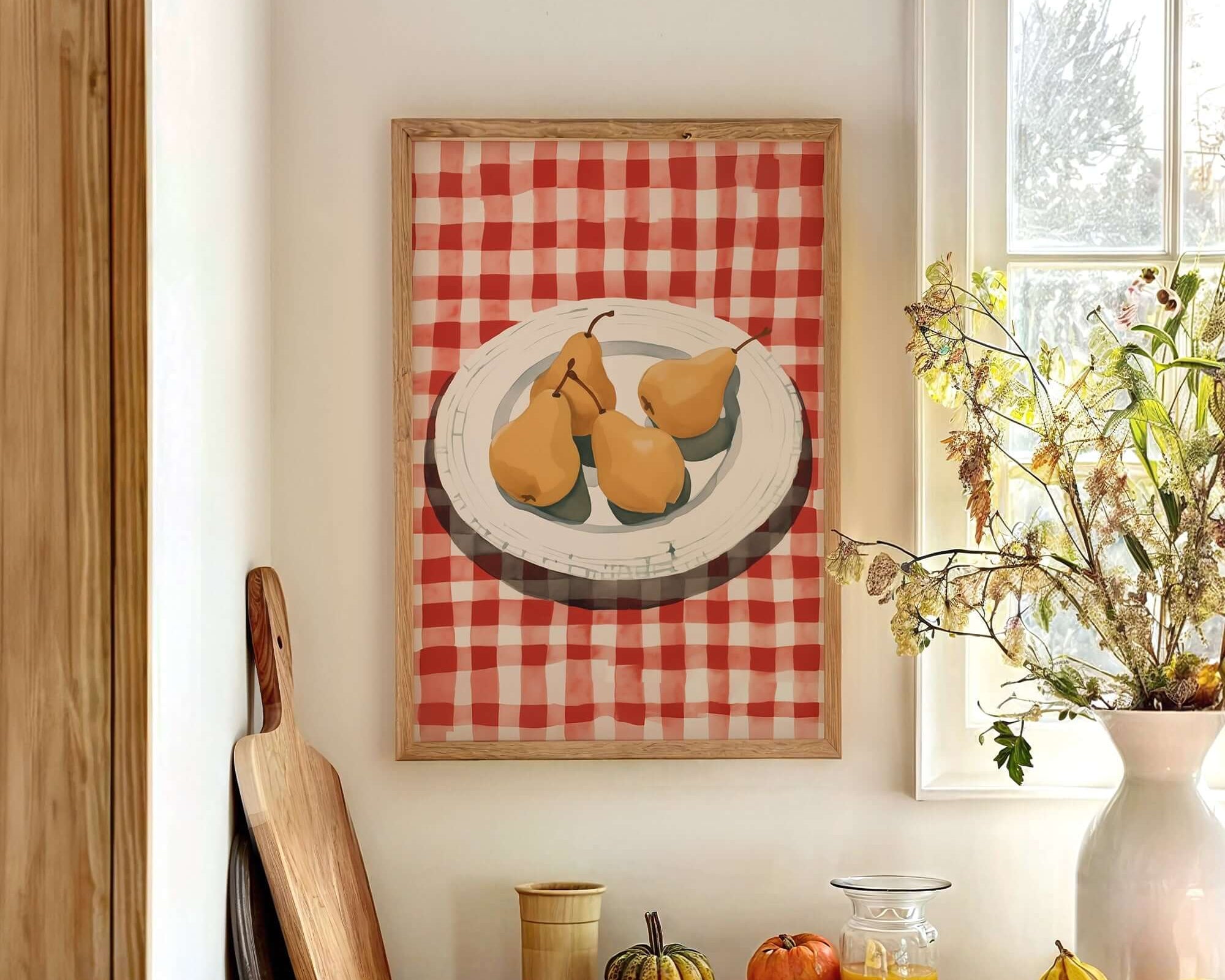 a kitchen counter with a framed painting of pears on a red and white checkered tablecloth, a wooden cutting board, a vase of flowers, and various fruits and vegetables on the counter.