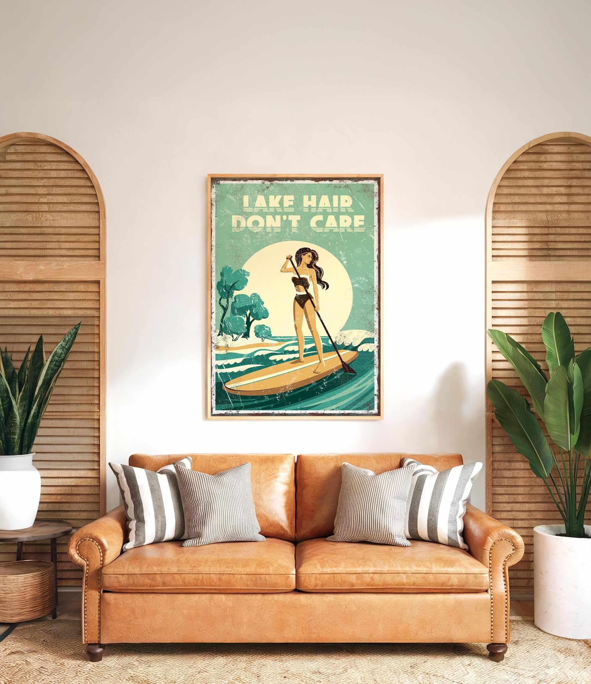 a vintage-style poster of a woman paddleboarding on a lake, with the text "LAKESHORE DON'T CARE" displayed prominently. The poster is hung on a wall above a brown leather couch, which has striped pillows on it. There are also two potted plants in the room, one on each side of the couch.