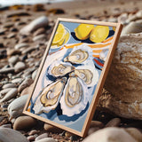 A framed painting of a seashell with a lemon slice on top, placed on a rocky beach.