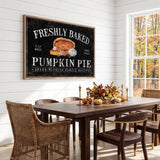 a dining room with a wooden table and chairs, a framed sign on the wall that reads "Freshly Baked Pumpkin Pie", and a vase of flowers on the table.