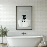 A framed poster with a black dog sitting in a bathtub, surrounded by bubbles, is displayed on a white tiled wall above a white freestanding bathtub.