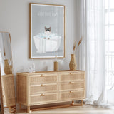 a wooden dresser with drawers, a framed poster of a cat in a bathtub, and a mirror on the wall.