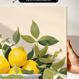 a hand holding a framed print of a still life composition with a bowl of lemons and green leaves.