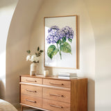 a wooden dresser with a framed painting of purple hydrangeas hanging on the wall above it. The dresser has six drawers and a vase of white flowers on top.