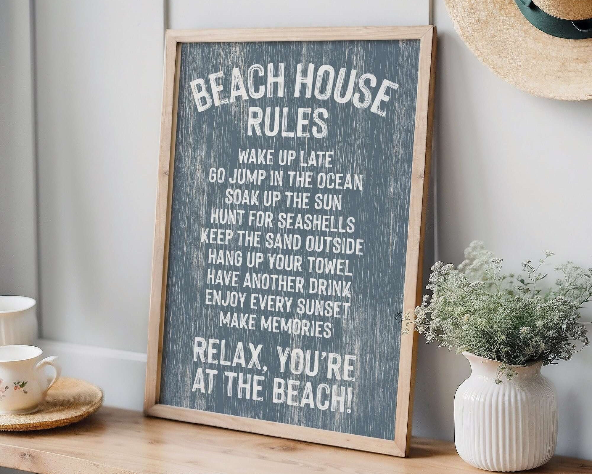 The Beach House Rules Sign, Painted Wood, Harbor Blue, sits on a wooden table beside a plant and straw hat—perfect coastal decor for adding charm to any space.