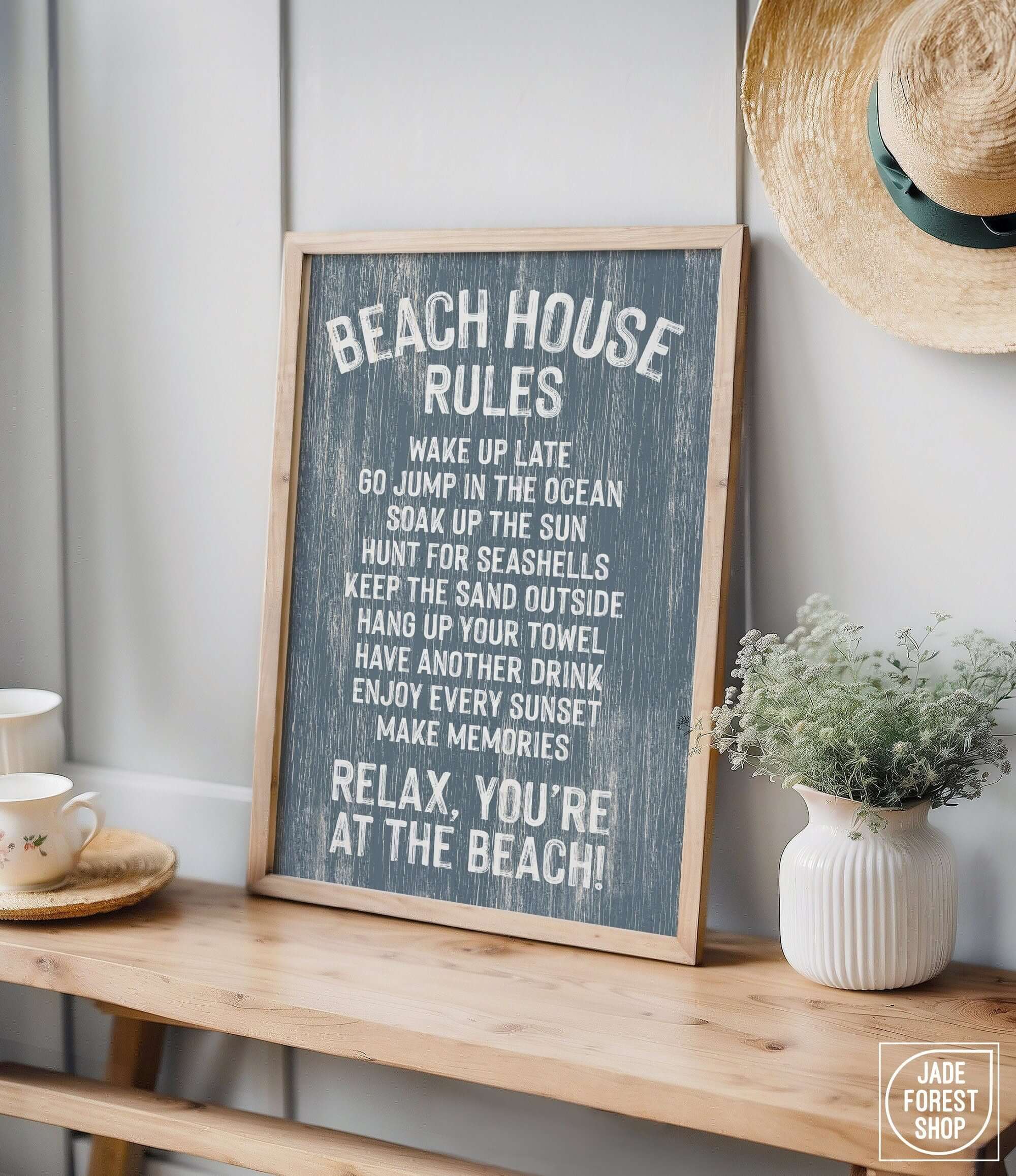 The Beach House Rules Sign, Painted Wood, Harbor Blue, sits on a wooden table beside a plant and straw hat—perfect coastal decor for adding charm to any space.