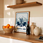 a kitchen counter with a framed poster of a mountain landscape with skis and Adirondack chairs,, a basket of oranges, a bottle of wine, and various decorative items such as vases and a bowl.