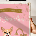 a hand holding a framed print with a pink background and a dog illustration. The text on the print reads "Wash Your Paws".