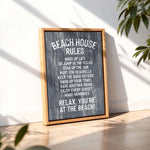 A framed sign with the text "BEACH HOUSE RULES" displayed on a wooden surface, with a plant in the background.