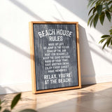 A framed sign with the text "BEACH HOUSE RULES" displayed on a wooden surface, with a plant in the background.