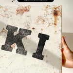 a close-up view of a white canvas with a large, bold black letter "K" prominently displayed. The canvas appears to be in a somewhat worn and aged condition, with visible rust and scratches.