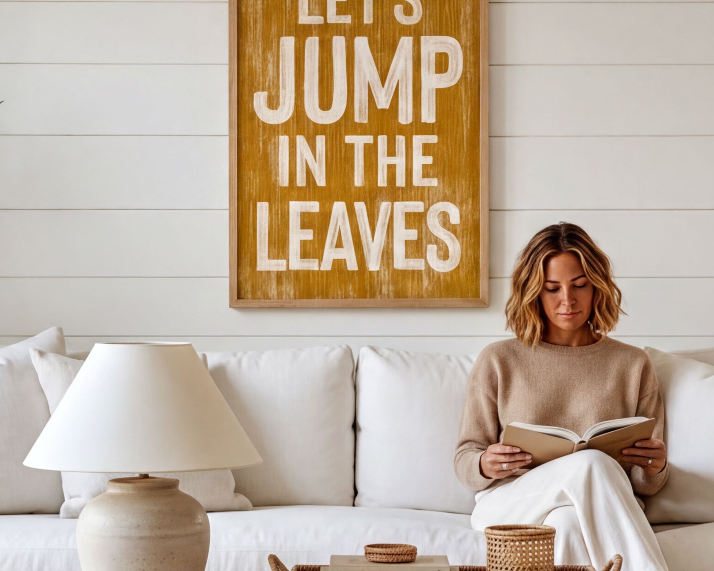 A woman is sitting on a white couch, reading a book, with a wooden sign above her that says "Let's JUMP in the LEAVES".
