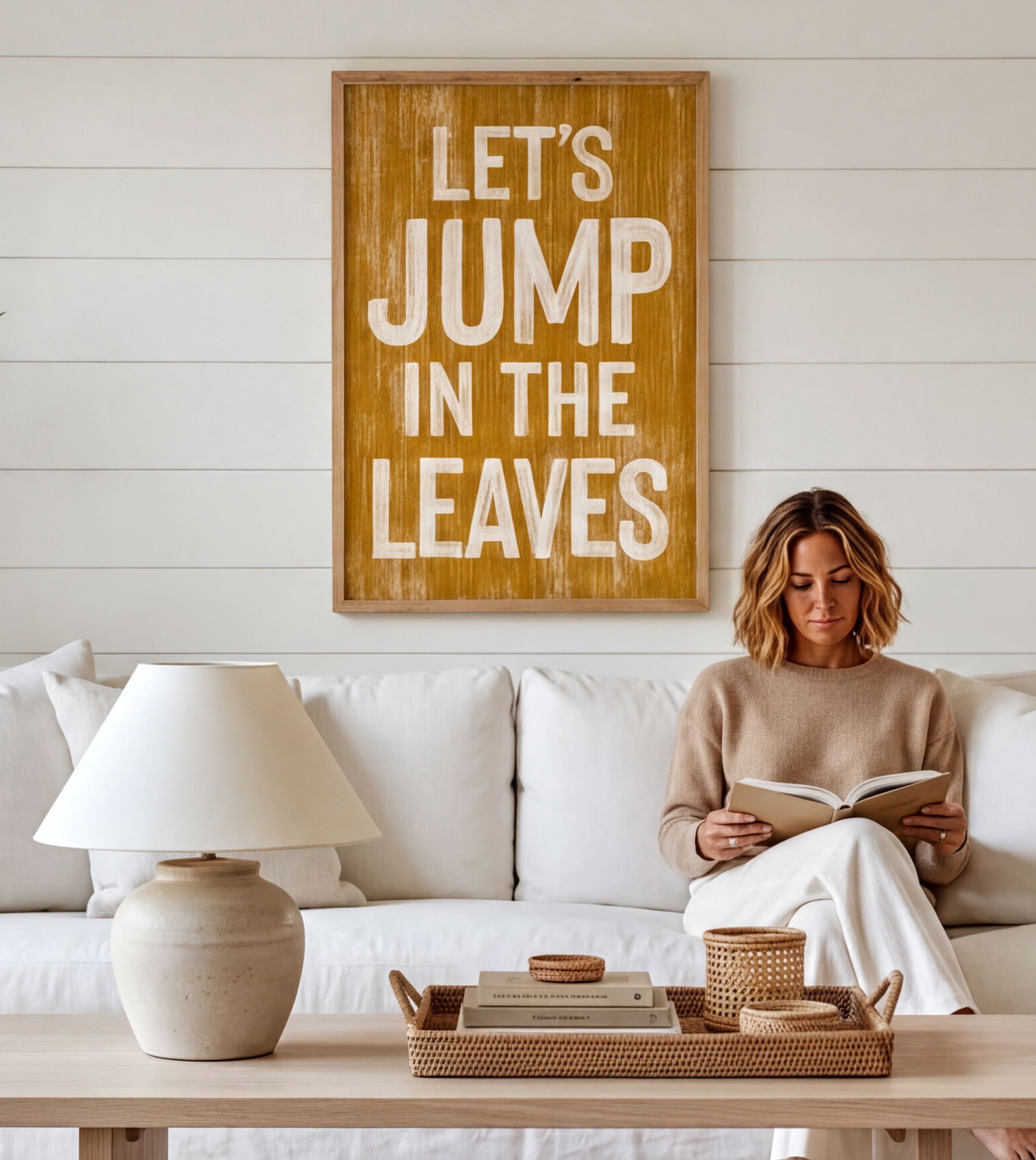 A woman is sitting on a white couch, reading a book, with a wooden sign above her that says "Let's JUMP in the LEAVES".