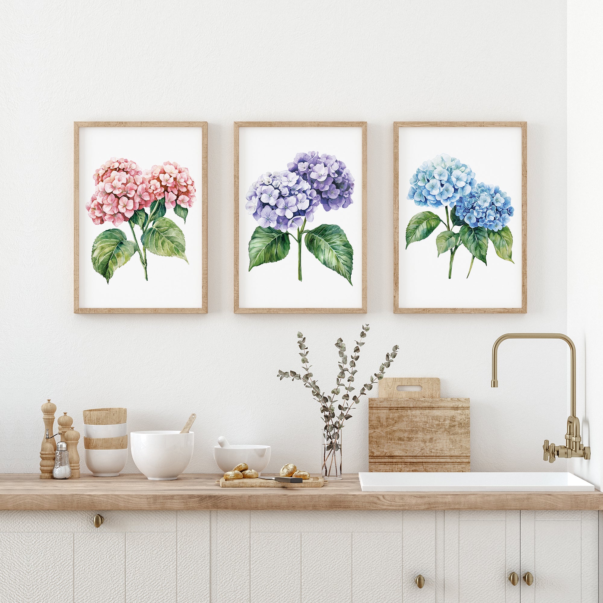 a kitchen counter with three framed watercolor paintings of hydrangea flowers hanging above it. The paintings depict different colors of hydrangeas - pink, purple, and blue. The counter also has various kitchen items, such as a bowl, a vase with flowers, and a sink.