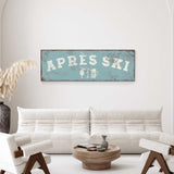 a living room with a white sofa, a wooden coffee table, and a decorative vase. Above the sofa, there is a vintage-style sign that says "APRES SKI" with a beer mug and a wine glass icon.