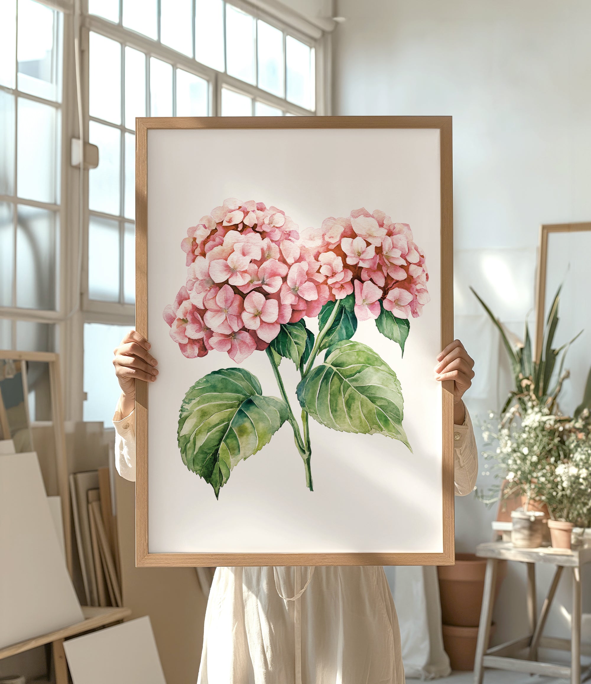 A person is holding up a framed painting of pink hydrangea flowers in a room with various potted plants and art supplies.