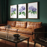 a living room with a brown leather couch, a wooden coffee table, and three framed paintings of purple hydrangeas hanging on the wall above the couch.