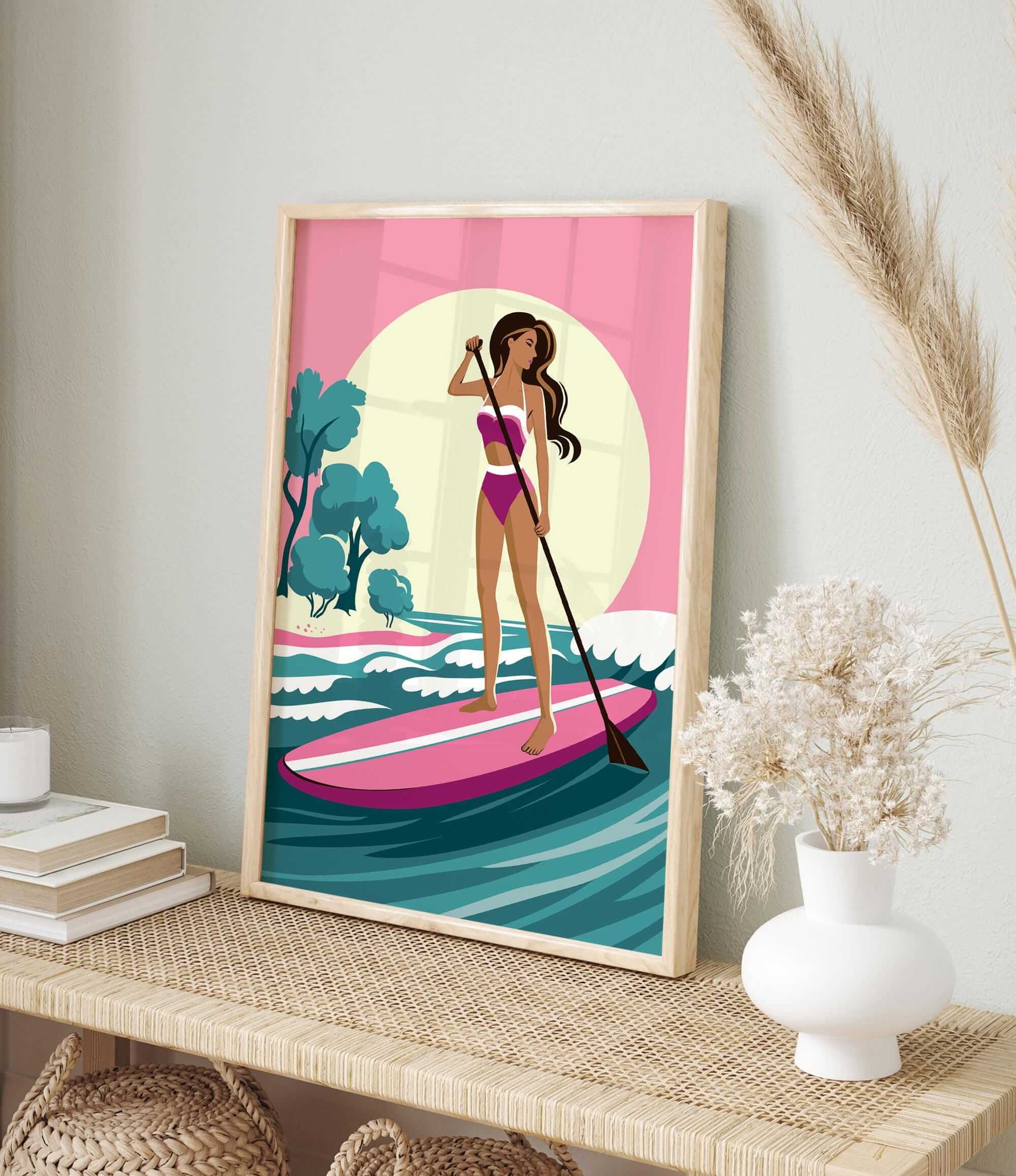 A framed illustration of a woman paddleboarding on a pink surfboard, with a pink and green background depicting a beach scene.
