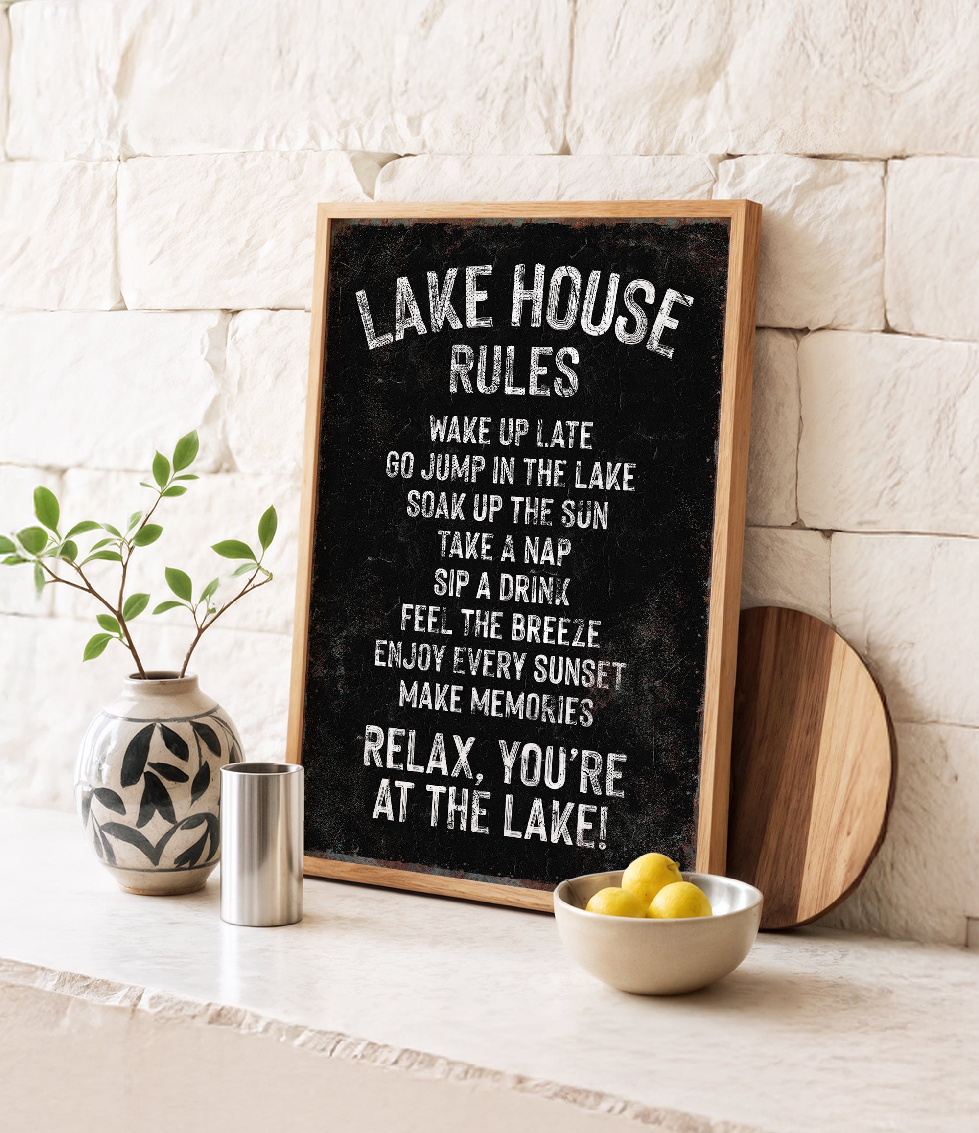 A blackboard sign with white text, a wooden board, a vase with a plant, a bowl of lemons, and a metal container on a white countertop.